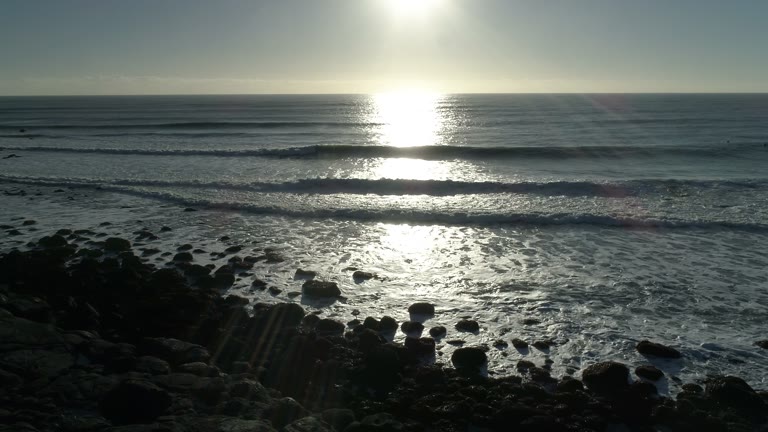Ocean waves roll onto a rocky coastline, with bright sunlight reflected across the water.